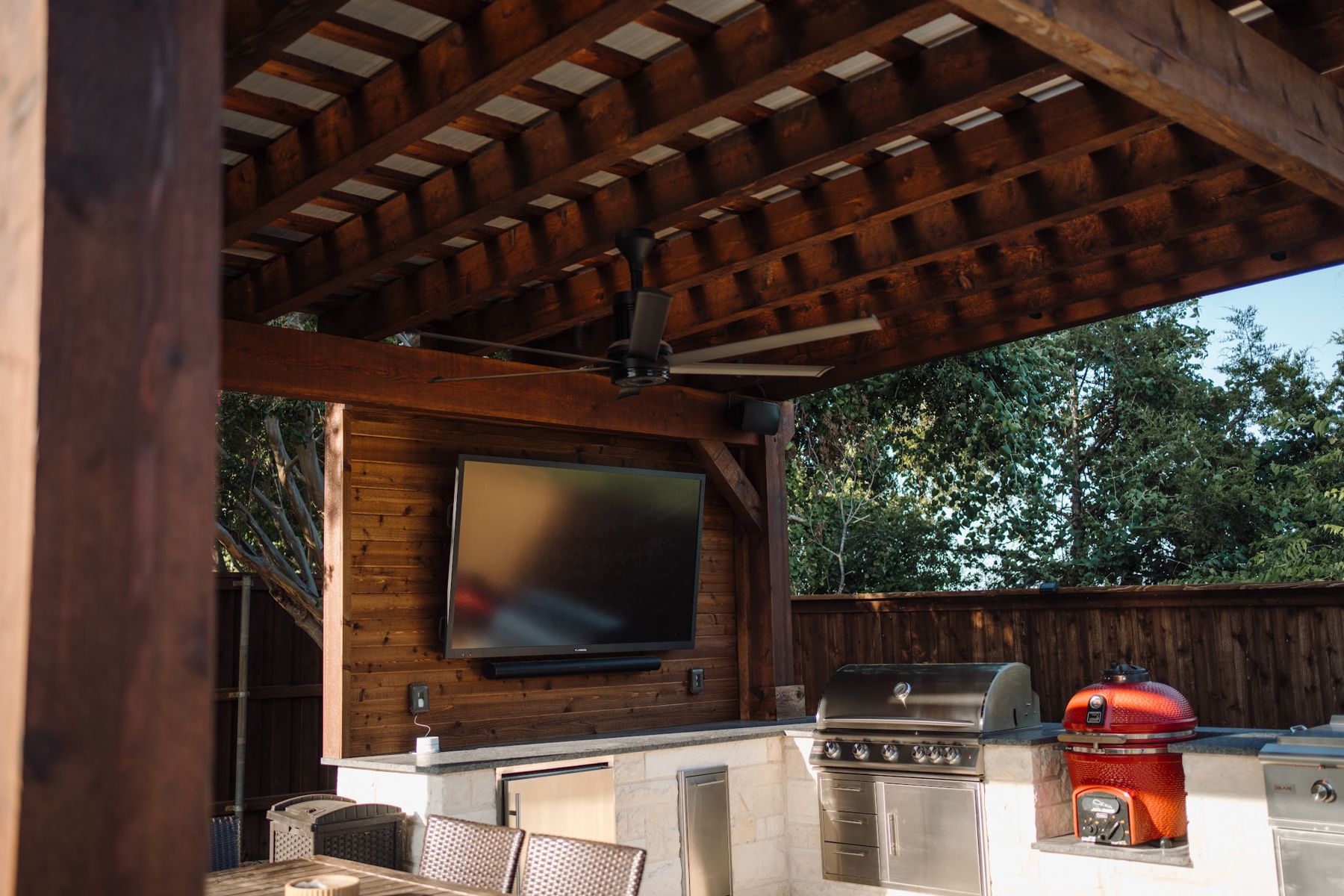 Cedar pergola over an outdoor kitchen with a built-in grill, red kamado cooker, stone island and wall-mounted TV in Benbrook