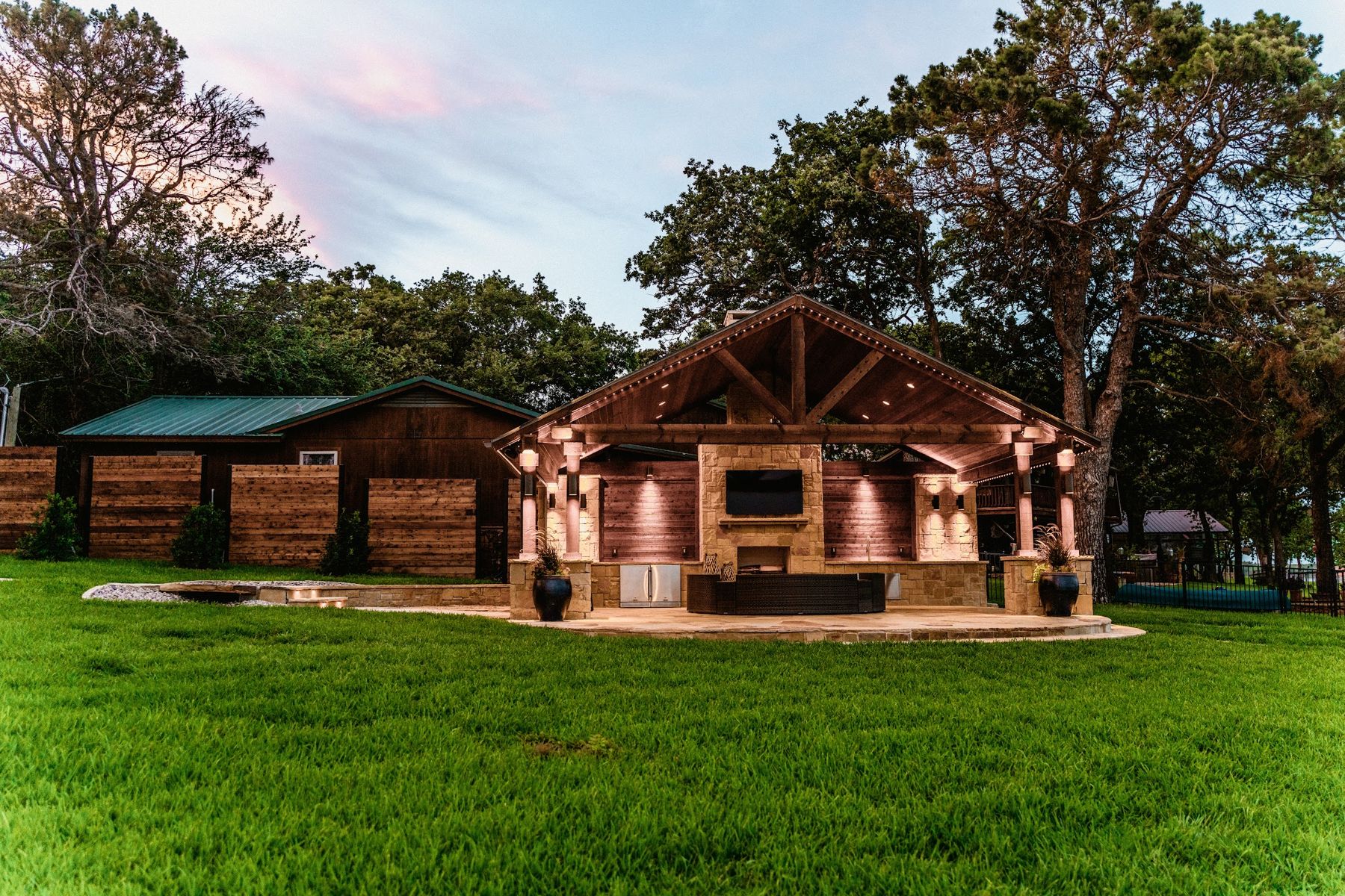 Timber pavilion with stone fireplace glowing at dusk on a lakefront lawn, Eagle Mountain Lake