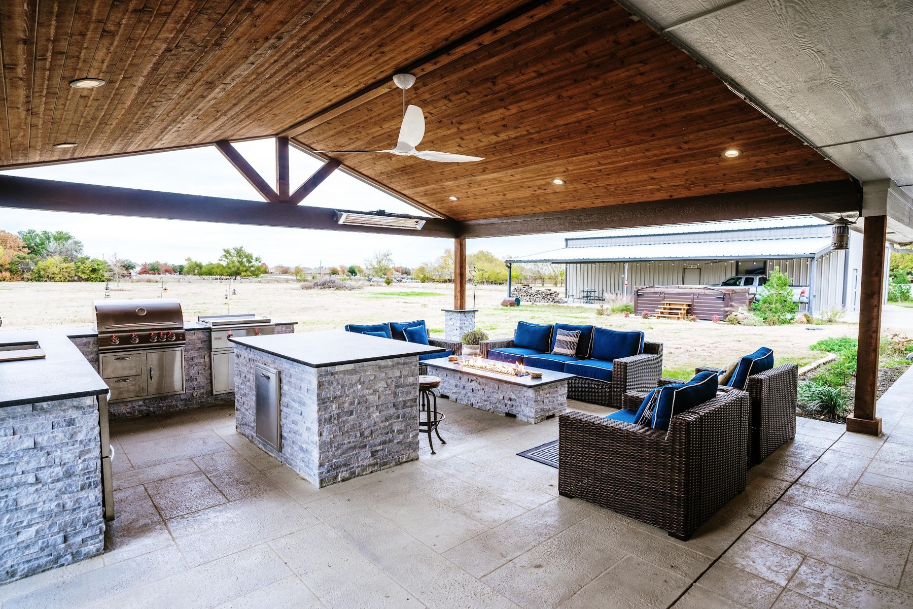 Covered outdoor kitchen with grill, island seating, fire table and blue lounge chairs under a cedar ceiling, Haslet