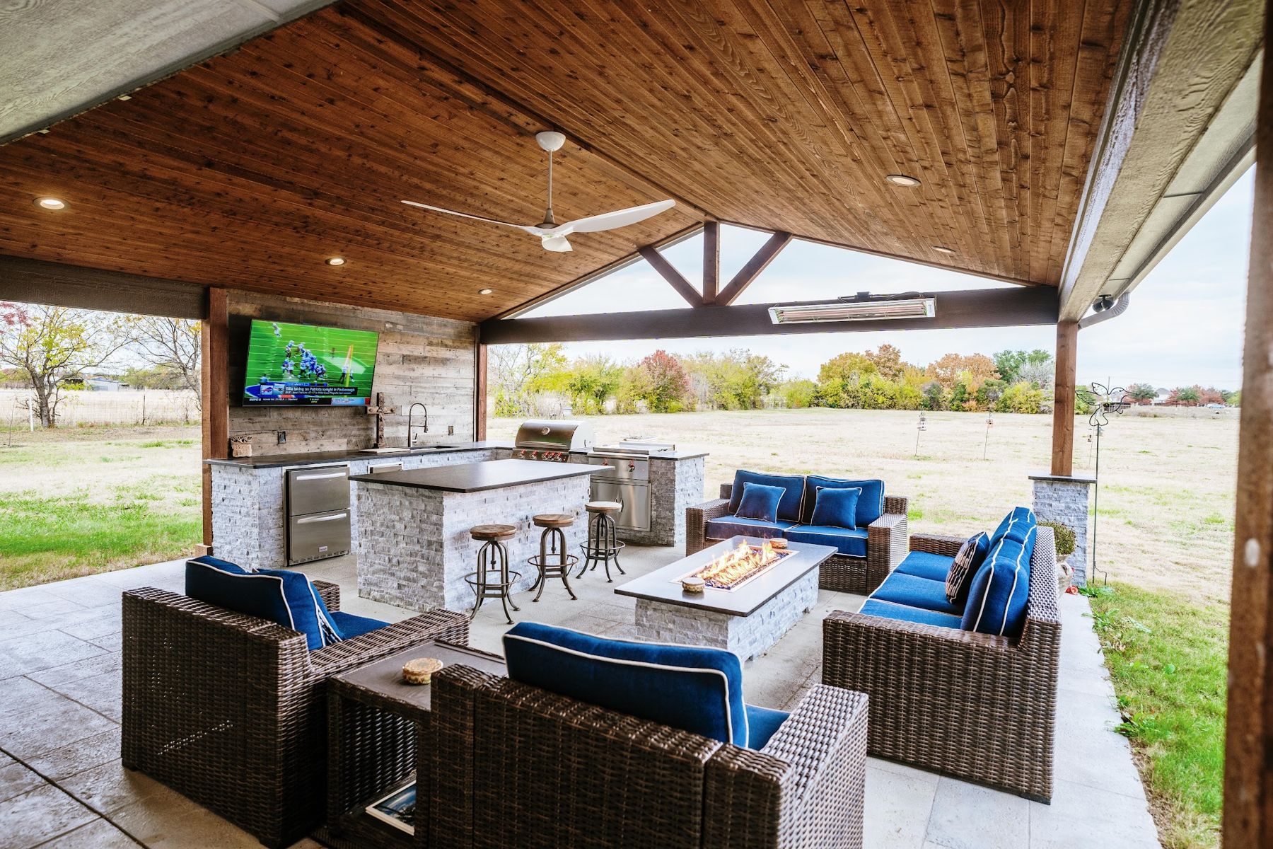 Covered patio with outdoor kitchen, TV wall, fire table and blue lounge seating, Haslet