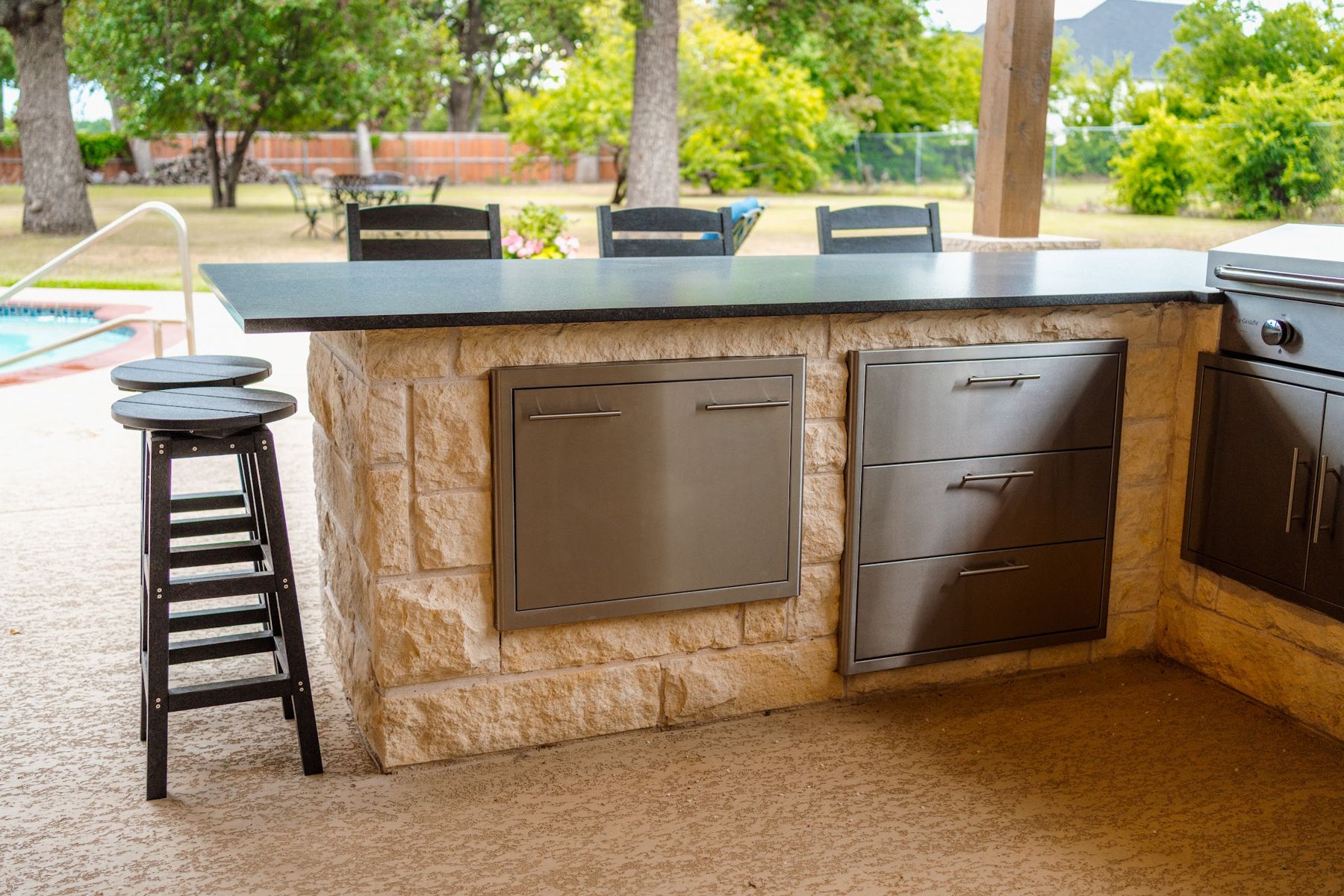Limestone outdoor kitchen with stainless doors and drawers beside a pool, Keller